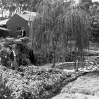 Image: Five women and two men pose for a photograph in a small terraced garden. A small pool in the middle of the garden is flanked by two trees and a stone wall