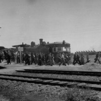 Image: A line of men and women walk away from a train towards a group of buildings in the background