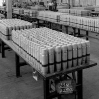 Image: A group of men and women stand in a large room full of rows of tables. Atop each table are several rows of artillery shells
