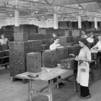 Image: A group of women and two men stand at tables in a large room and manufacture rectangular metal containers