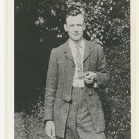 Bespectacled man in two piece suit and tie holding a pipe, and standing in a garden