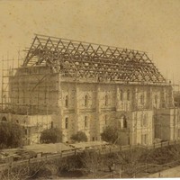 Image: A large hall is under construction. The stone walls are covered in scaffolding and the hammerbeams of the roof are visible. Men can be seen on the roof of the building.