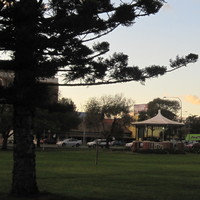 Image: A grassy park interspersed with trees and a single wooden gazebo. A large road with several cars and a number of large buildings are visible in the background