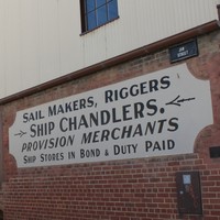 Image: A two-storey building with brick first floor and corrugated metal second floor. The words ‘Sail Makers, Riggers, Ship Chandlers. Provision Merchants Ship Stores in Bond Duty Paid’ are painted on a sign attached to the building’s first floor 