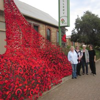 Image: three women standing next to large number of crocheted poppies draped from building