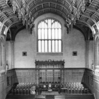 Image: the interior of a large hall with stone and wood panneled walls and a decorative wooden ceiling. The rear wall features a large window underneath which is a stage area with rows of chairs upon it and more chairs facing it. 