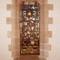 Image: a stained glass window featuring three children, one playing a lute and one which appears to have wings, and a lamb gathered around a man reading a book.