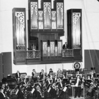 Image: an orchestra plays on a stage in front of a large pipe organ. The organ has two balcony areas within it upon which stand a man and a woman in formal dress.