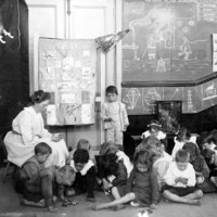 Image: a caucasian woman sits on a stool surrounded by children in a classroom with blackboards on the walls