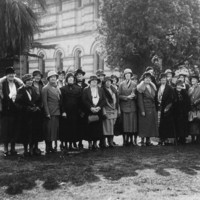 Image: A group of women standing in a line in front of two trees and a building, they all wear hats and long skirts with coats