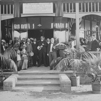 Image: a group of men and women in early 20th century dress sit or stand in front of a doorway in the shade of a verandah decorated by flags. Palms in pots next to the stairs leading to the verandah dominate the foreground.