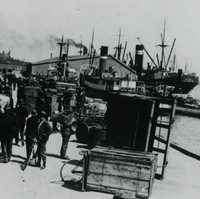 Image: A group of men mill about on a wharf. Two steamships are visible in the background. What appears to be a horse-drawn cart tipped on its side is located at the edge of the wharf