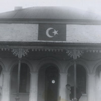 Image: An Islamic symbol of a crescent moon and star is painted just below the roof on a building fronted with arches. A man bends down behind a narrow column with one leg bent at the knee, possibly removing or putting on a shoe.