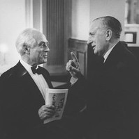 Image: Two men in tuxedos stand face to face. The man on the right has his hand raised, one finger pointed up, gesturing. The other, who holds a piece of paper in his hand, is smiling.