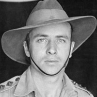 Image: An upper body portrait photograph of a man in an army uniform, and wearing an army hat with chin strap. 