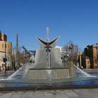 Image: grey three pointed fountain with city buildings in background