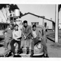 Image: four young men sitting on bonnet of car, buildings in background