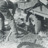 Image: A young Caucasian man in utility clothing uses a vehicle jack to attempt to extricate the bogged down rear wheel of a 1930s vintage utility truck