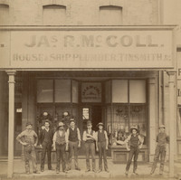Image: A group of Caucasian men in late nineteenth-century labourer attire stand in front of a shop. The words ‘Jas. R. McColl, House and Ship Plumber, Tinsmith, &c.’ are painted on the front of the building