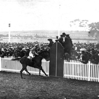 Image: Black and white photo of horseracing in progress, crowd behind a fence