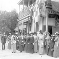 Image: A group of women and men in Edwardian attire stand in front of a two-storey stone building with flags hanging from the second-floor verandah
