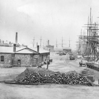 Image: Several late nineteenth century sailing ships are moored alongside a large wharf in a port town. A large pile of stone ballast and rail cars are visible in the foreground, while a single-storey bluestone building is located nearby