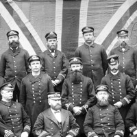 Image: A group of men attired in nineteenth-century South Australian customs service uniforms pose for a photograph. A large Union Jack flag appears as a backdrop behind the men