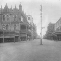 Image: a wide intersection with a lamppost in the centre. On one corner is an ornate three storey building with a verandah, balcony and highly decorative and detailed parapet.