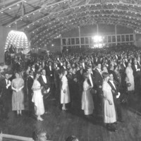 Image: a crowd of men and women wearing 1920s era clothing stand in pairs in a large hall the ceiling of which is decorated with lines of small lights. 