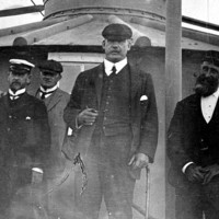 Image: A group of middle-aged Caucasian men in early Edwardian attire pose for a photograph outside a metal lighthouse. One man is wearing what appears to be a government uniform