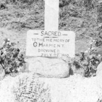 Image: Two middle-aged Caucasian men in Edwardian attire flank a simple grave site. The grave is covered with stone and features a large white wooden cross, to which is attached a sign that reads ‘Sacred to the memory of G. Marment, drowned July 28 1910’