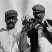 Image: Two young moustachioed Caucasian men in Edwardian-era work clothing pose for a photograph. The men stand in front of a cottage and white picket fence, and the man on the left holds a large crayfish in his left hand