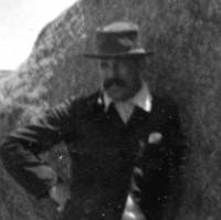 Image: A young moustachioed Caucasian man in casual early Edwardian attire poses for a photograph next to a large rock formation on an island