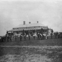 Image: Black and white photograph of a gathering of people. Some are standing on the verandah of a house. Another groups stands further back from the house (closer the camera) in a large circle, joined by their hands