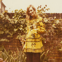 Image: A woman posing for a photograph outside. Behind her is a brick wall covered in vines. She is holding a yellow flower and smiling at the camera.