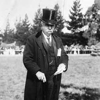 Image: A middle-aged man wearing a suit with top hat and holding a cane stands in a paddock. A fence and buildings are visible in the background