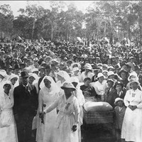 Image: Black and white photograph of a large crowd standing behind a group of women dressed in white and a man in a dark suit. In the centre of the picture a wounded soldier can be seen in a stretcher bed. 