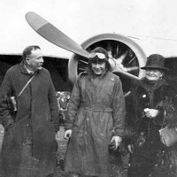 Image: A man in First World War-era aviator attire stands in front of an aircraft with two men and two women