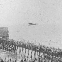 Image: A large crowd of people watch an early twentieth century aircraft make a low pass over a jetty near a seaside community