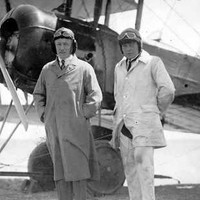 Image: Two men wearing suits, overcoats and early-twentieth century aviator helmets pose for a photograph in front of a biplane