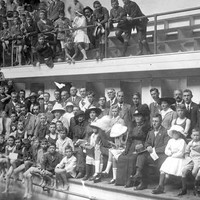 Image: A crowd of people in early twentieth century attire are gathered at the side of a large swimming pool