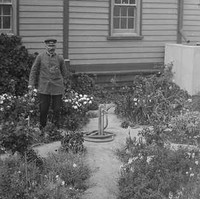 Image: A man in uniform stands in a flower garden next to a cottage
