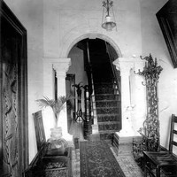 Image: the entry hall of a house. The decorative tiled floors are covered by a runner carpet which extends through an archway supported by columns and up a narrow staircase. 