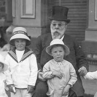 Image: Black and white photograph of seated man, wearing a top hat, surrounded by four small children. In the background a woman stands near a table facing away from the camera. 