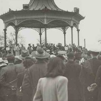 Image: a crowd of people, some in military uniform, watch dignitaries give speeches from a rotunda. A flag, possibly the Union Jack, is just visible on a flagpole to the right of the photograph. 