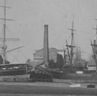 Image: A multi-storey stone building with large brick chimney sits near a river-front wharf. A larger chimney is located to the right of the building, and several ships are moored at the wharf