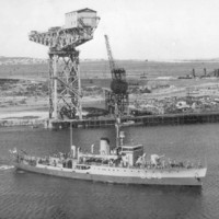 A warship gets underway in waters a short distance from a shore-based shipbuilding facility. The facility includes two large cranes and several buildings