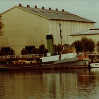 Image: A derelict tugboat is moored against a wharf next to a large complex of buildings. A number of other small boats are also moored at the same wharf