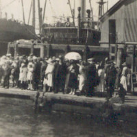 Image: A large group of people congregate on a floating dock next to a wharf. A steamship and warehouses are visible in the near background