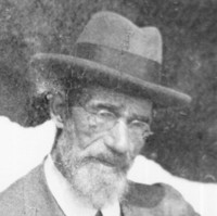 Image: A photographic portrait of an elderly Caucasian man wearing an Edwardian-era suit and hat. He has a white full beard and is wearing wire-rimmed spectacles